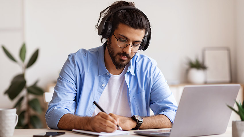Person sitting at a desk writing on paper next to a laptop